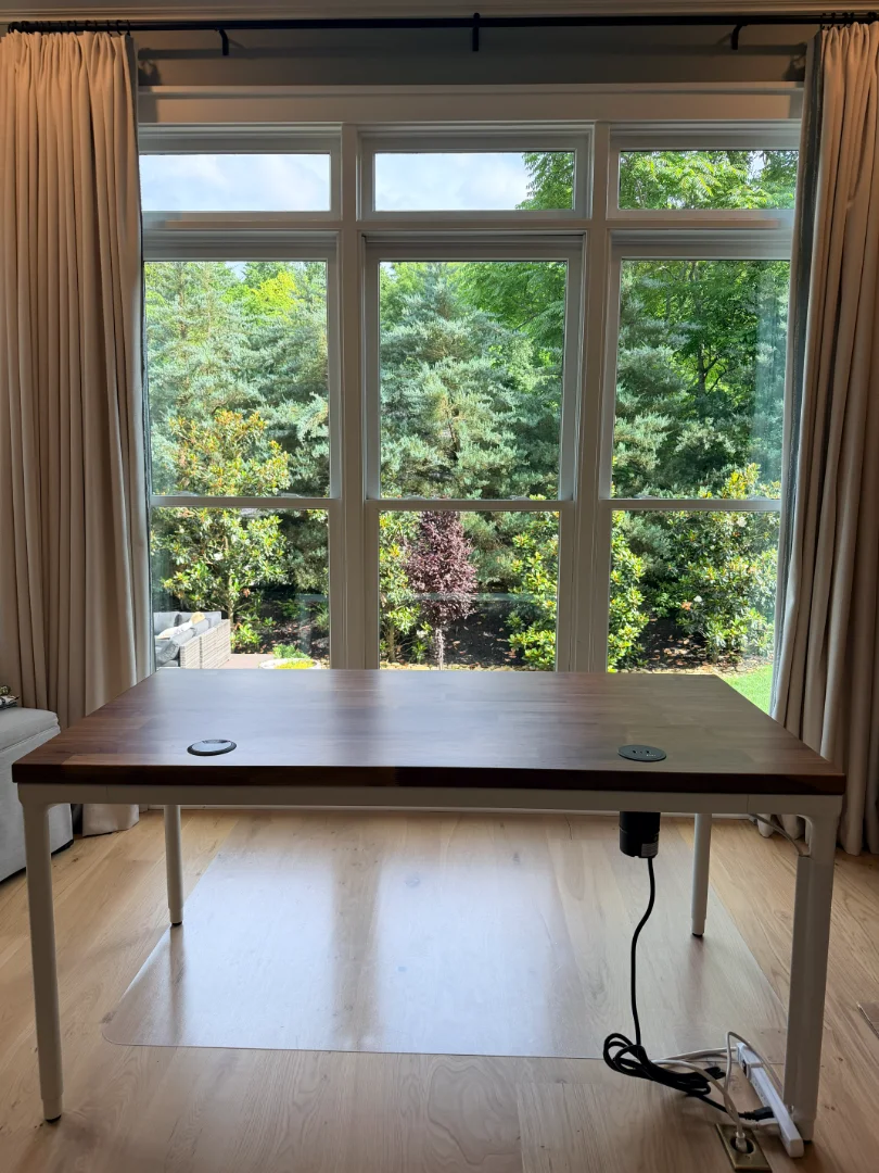 A fully assembled UPLIFT Desk Parsons Standing Desk with a wood tabletop and white frame positioned in front of large windows with curtains and a garden view.